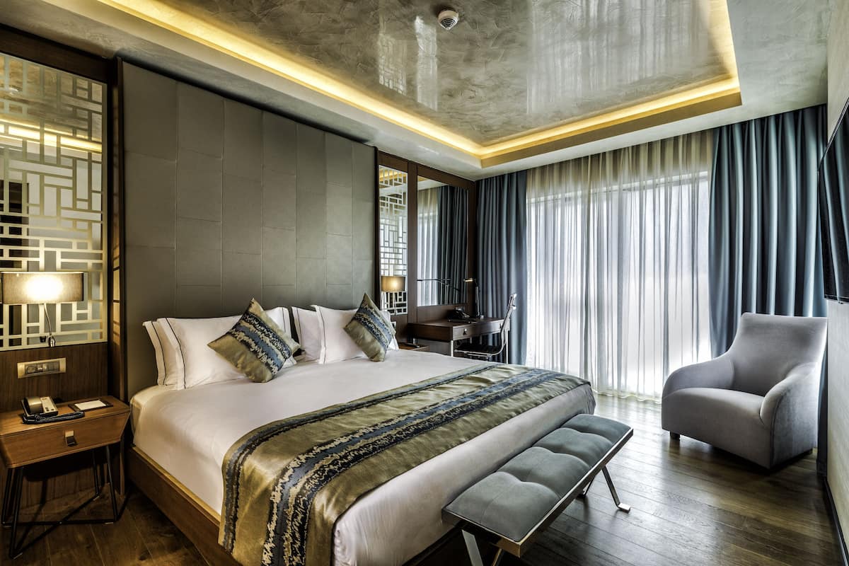 A modern hotel room with a large bed, a desk, a chair, and a view of the outdoors.