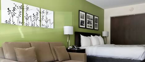 A hotel room with a beige sofa, a wooden coffee table, a bed with white bedding, a nightstand with a lamp, and wall art.