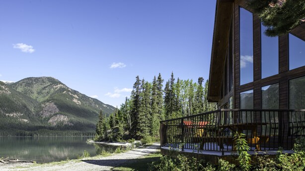 A lakeside cabin with a deck overlooking a calm lake and forested mountains. - Northern Rockies Lodge (Muncho Lake)