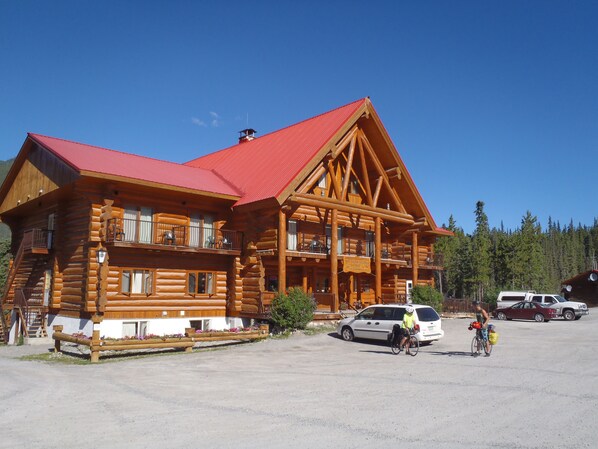 A large log cabin with a red roof, multiple balconies, and a spacious parking area.