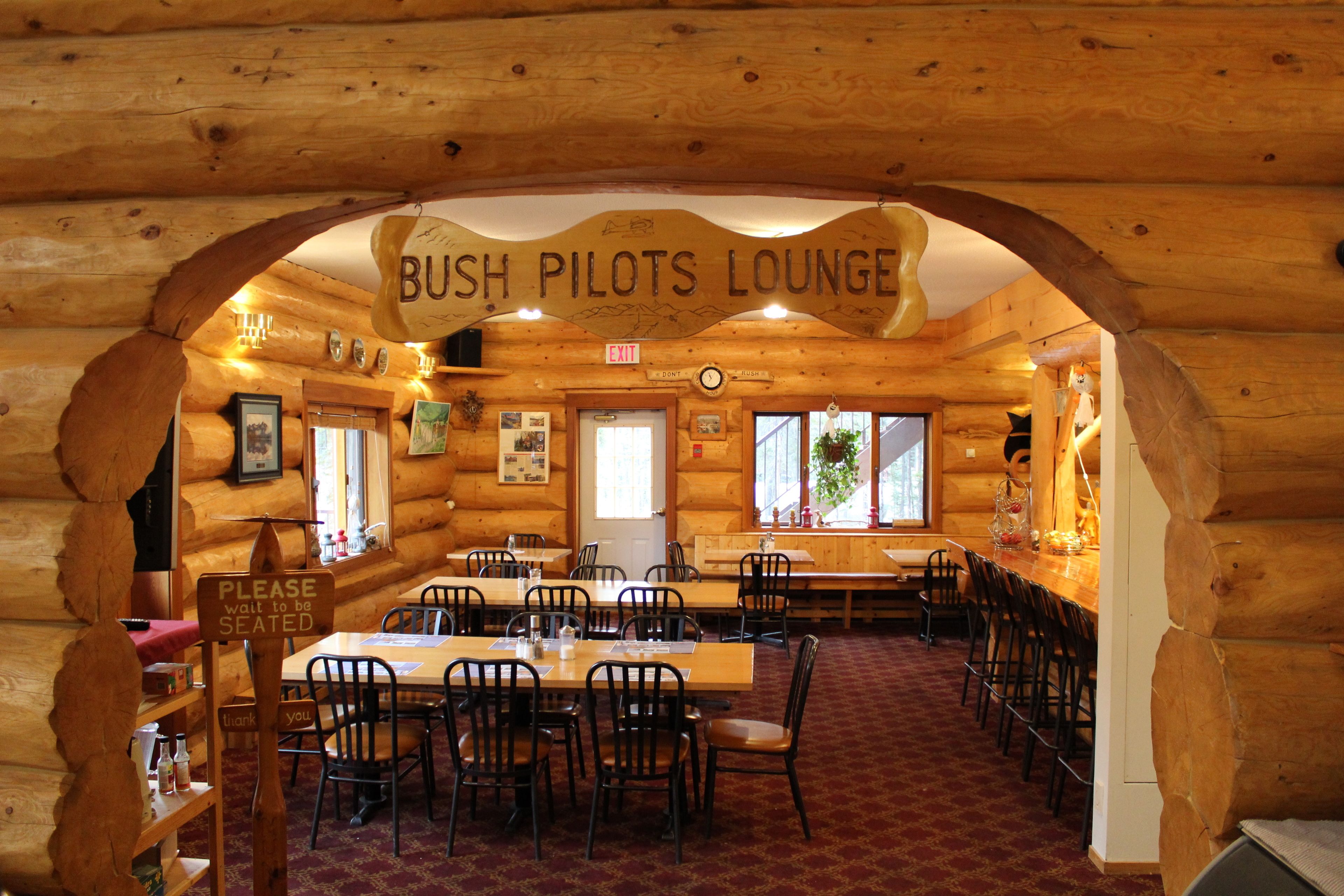 A log cabin interior with a 'Bush Pilots Lounge' sign, wooden walls, tables, chairs, and a carpeted floor.