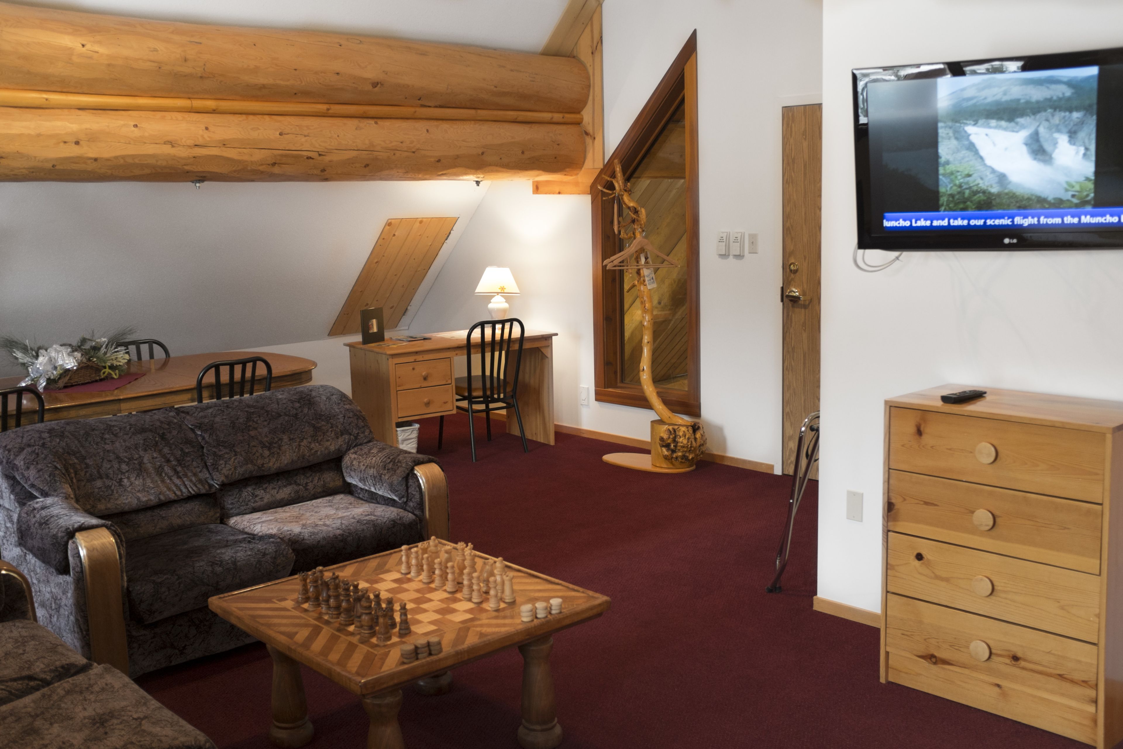 A cozy living room with a wooden ceiling, a television mounted on the wall, a chess table, a sofa, and a wooden cabinet.