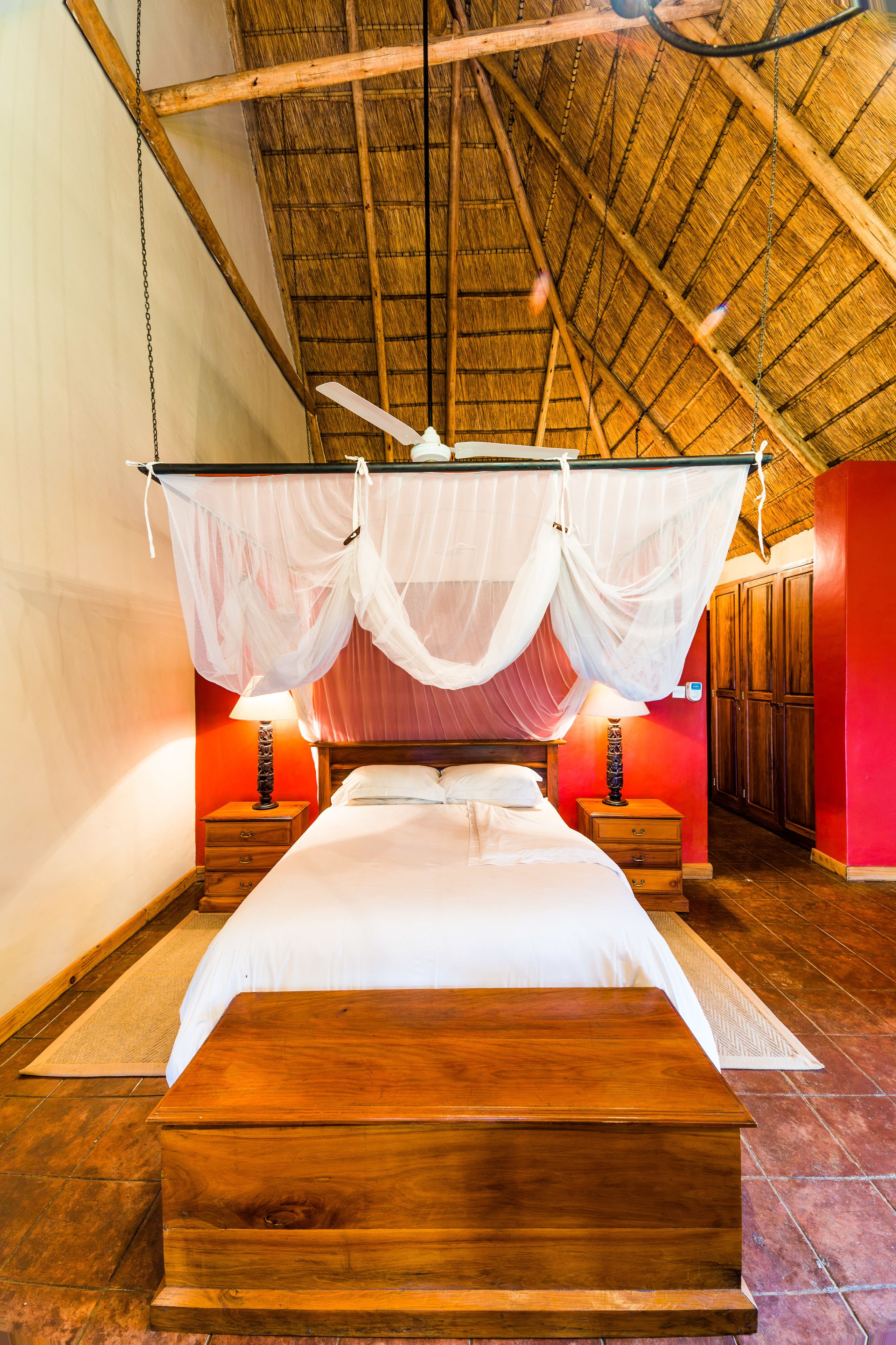 Una habitación con una cama bajo un tejado de paja, mesitas de noche de madera y una pared roja.