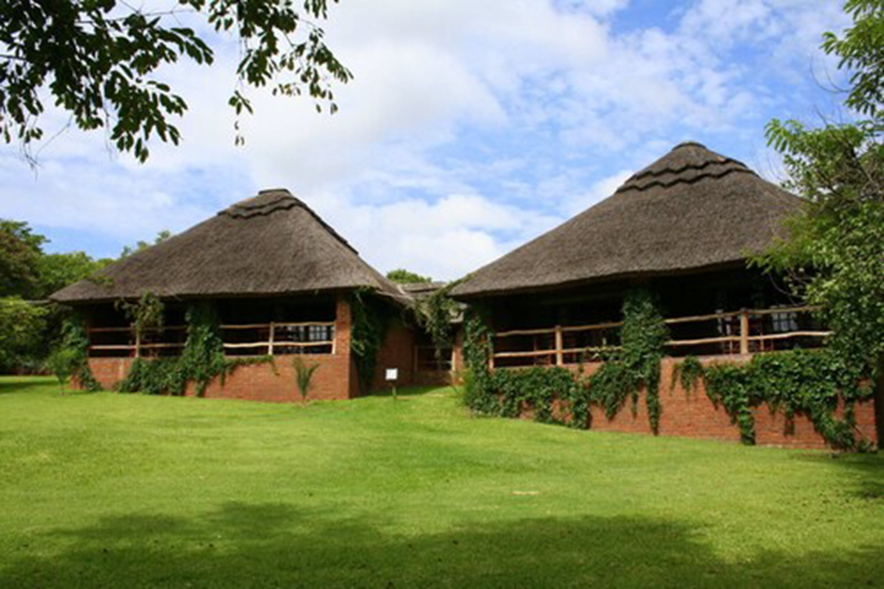 Two thatched-roof huts with brick walls and wooden railings, surrounded by a grassy area and trees.