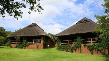 Two thatched-roof huts with brick walls and wooden railings, surrounded by a grassy area and trees.