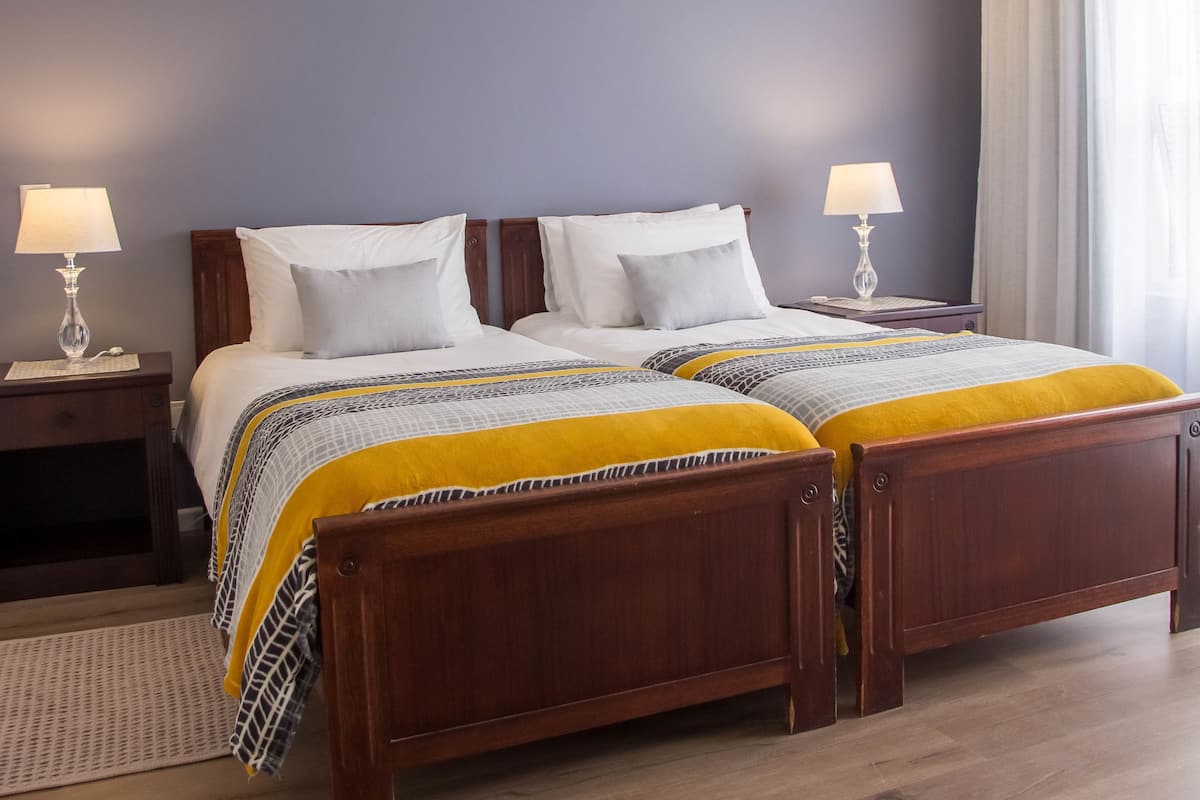 Una cama doble con estructura de madera, dos mesitas de noche con lámparas, un ventilador de techo y un ventanal con cortinas.