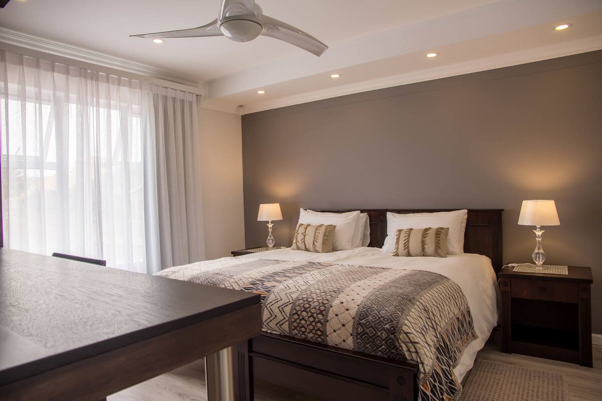 Una habitación de hotel moderna con una cama grande, dos mesitas de noche con lámparas, un ventilador de techo y un ventanal con cortinas translúcidas.