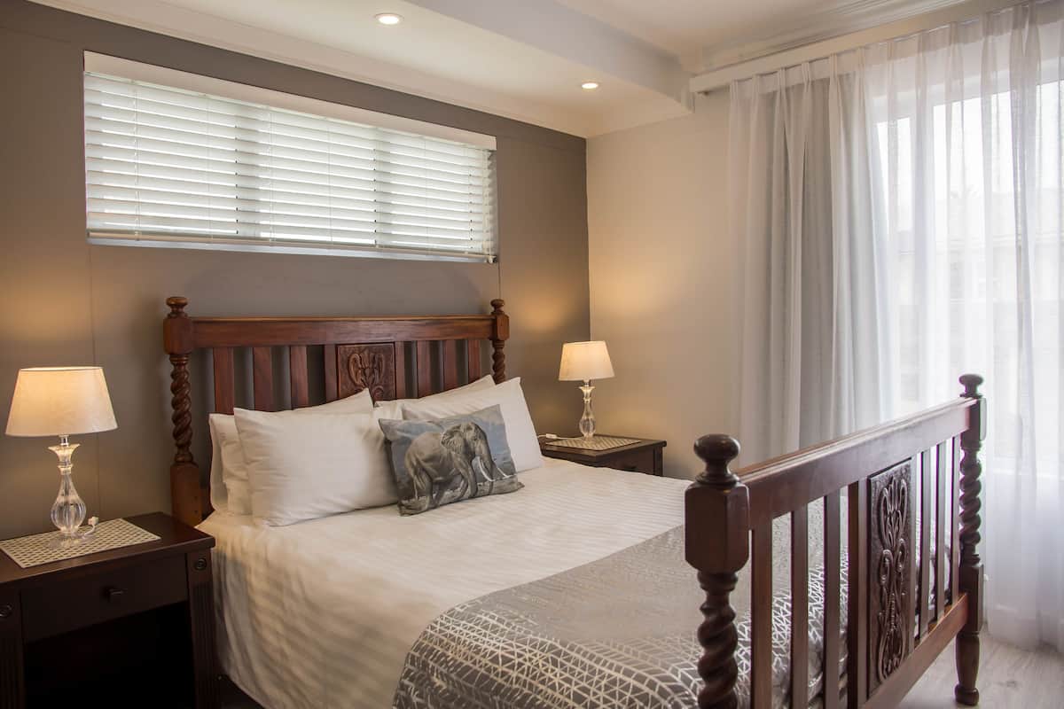 Una cama bien tendida con un cabecero de madera, dos lámparas de noche y una ventana con persianas.