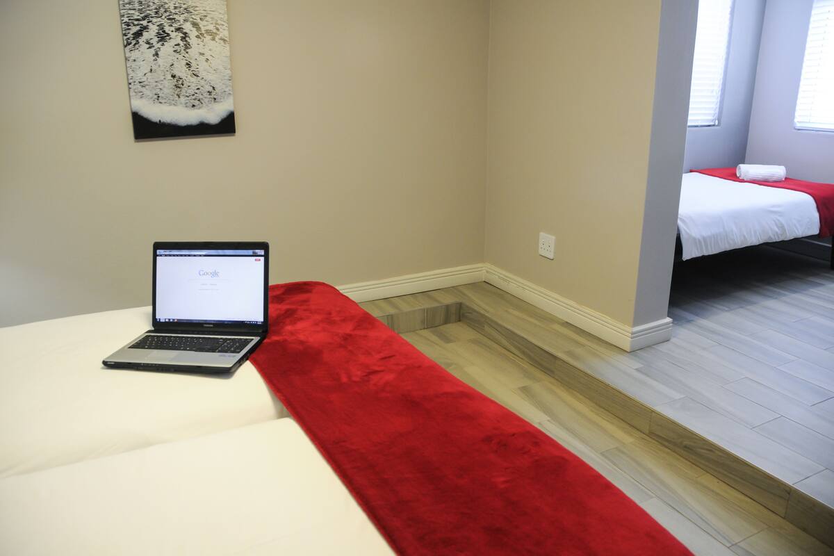 Una habitación de hotel con una cama, una laptop sobre la cama y un corredor rojo.