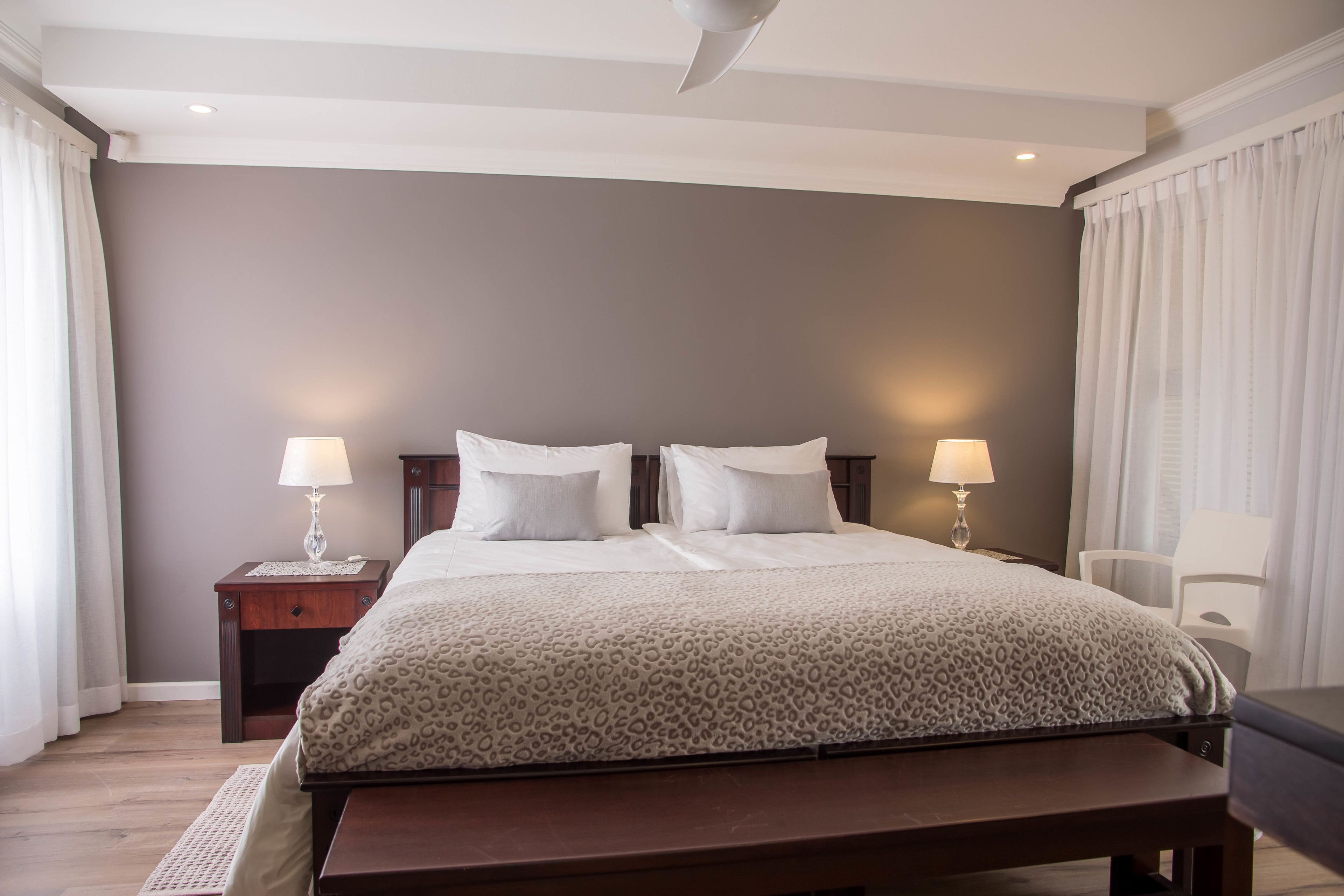 Un dormitorio con una cama grande, dos mesitas de noche con lámparas, un banco al pie de la cama y una ventana con cortinas.