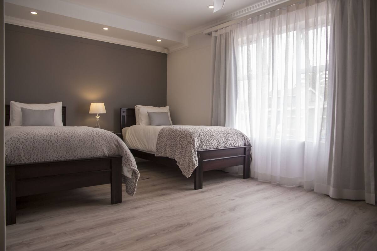 Un dormitorio con dos camas, una mesita de noche con lámpara y cortinas translúcidas.