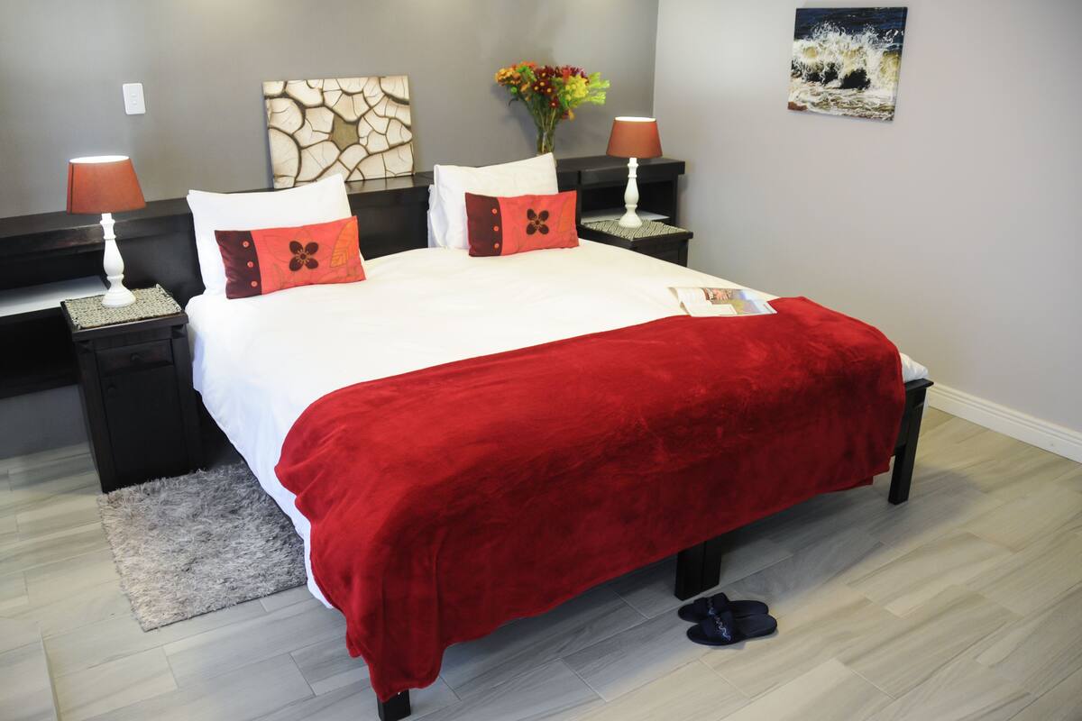 Una cama bien hecha con una colcha roja, dos mesitas de noche con lámparas, un florero y cuadros en las paredes.