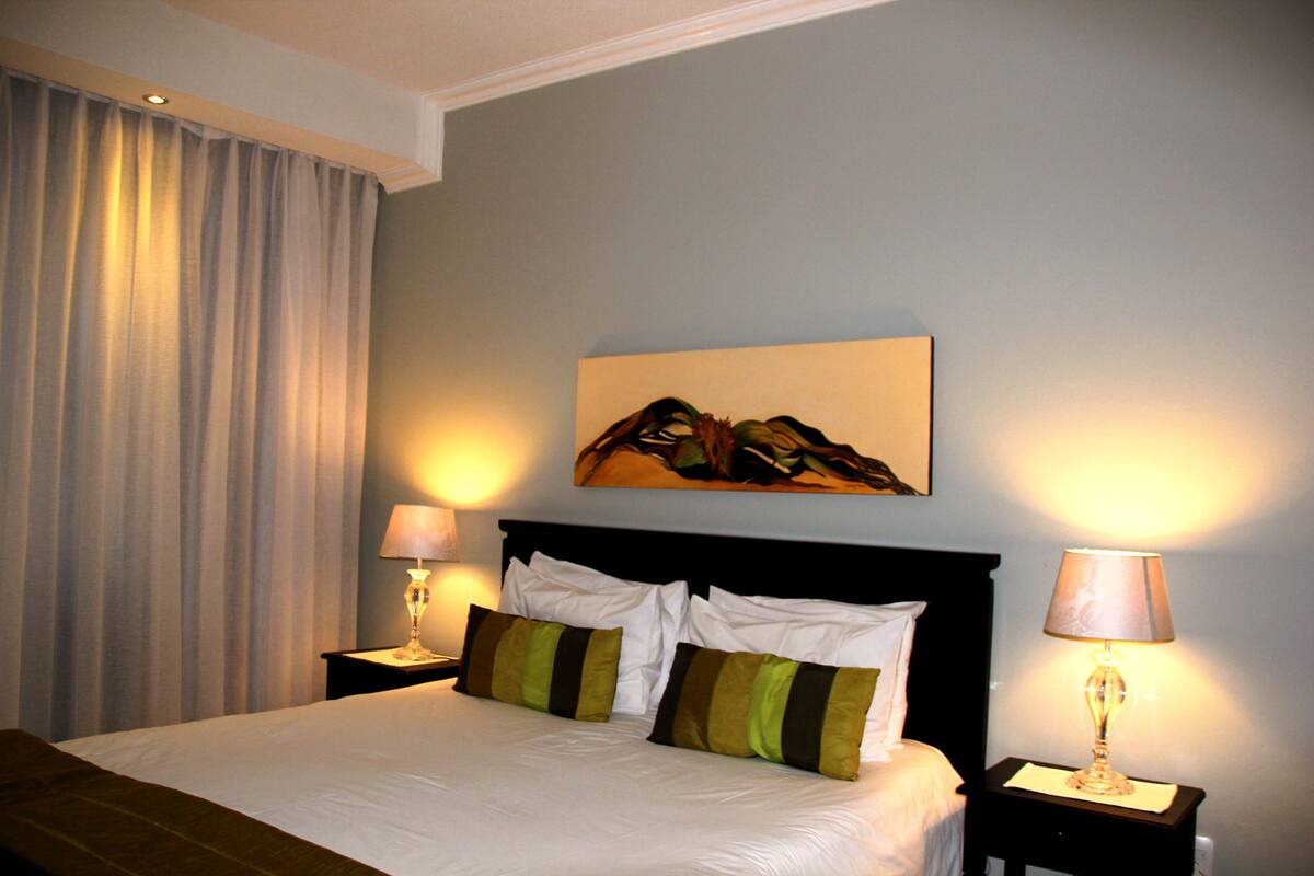 Una cama bien hecha con sábanas blancas y dos almohadas, flanqueada por dos mesitas de noche con lámparas, en una habitación con un ventanal grande y un cuadro en la pared.