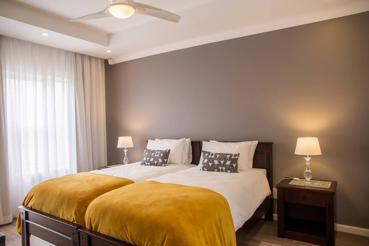 Una cama doble con colcha color mostaza, dos lámparas de noche y un ventilador de techo.