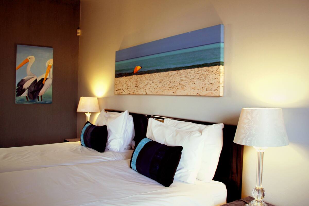 Habitación de hotel con una cama grande, dos lámparas fijadas a la pared y dos cuadros que representan una playa y pelícanos.