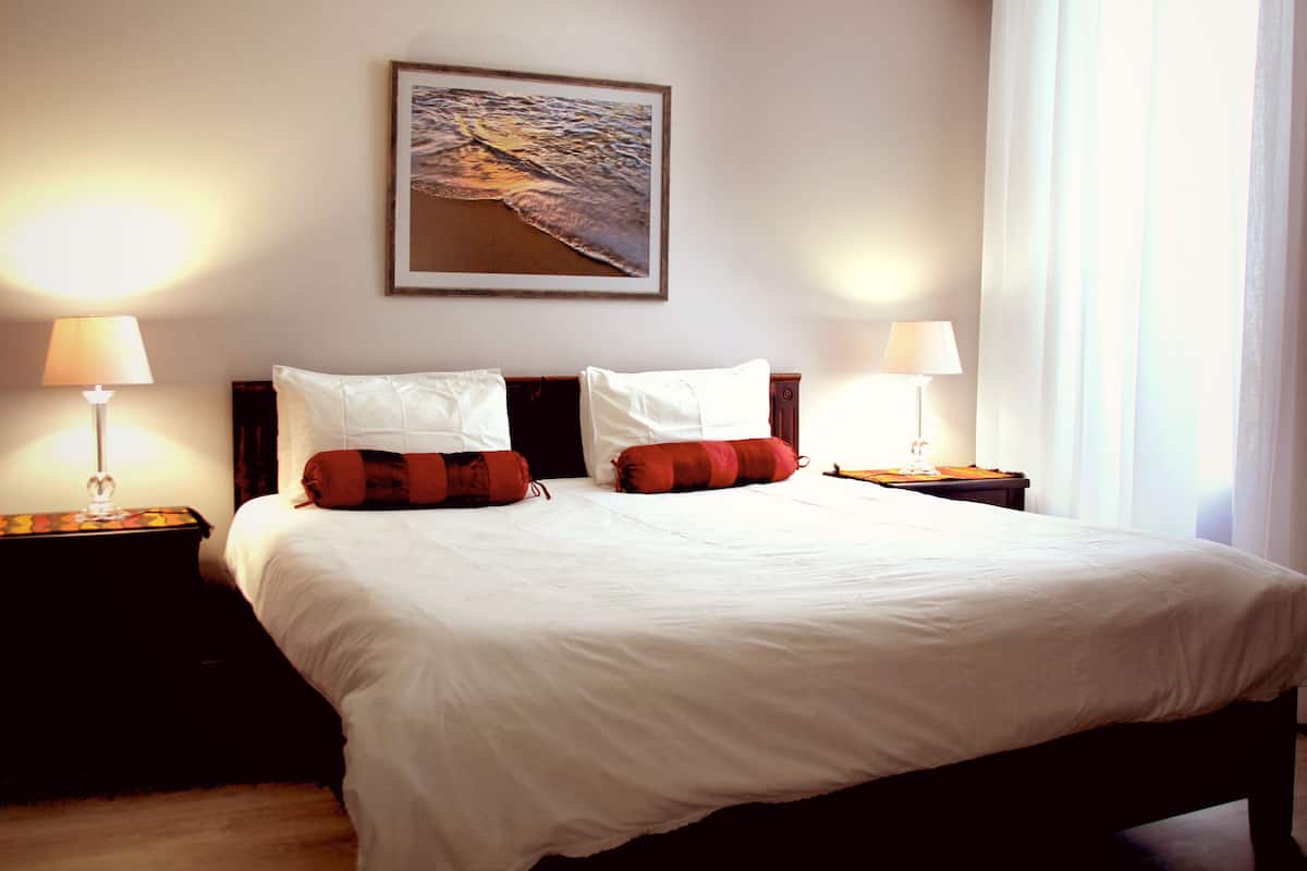 Una cama bien hecha con sábanas blancas, dos almohadas y una almohada a rayas marrón y roja. Hay dos mesitas de noche con lámparas, un cuadro en la pared y una ventana con cortinas.