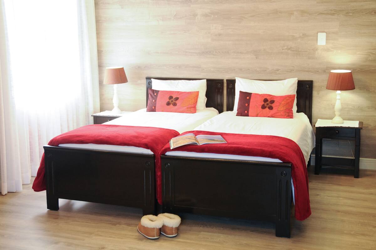 Una cama doble con una colcha y almohadas rojas, una mesita de noche con una lámpara y un par de zapatillas en el suelo.
