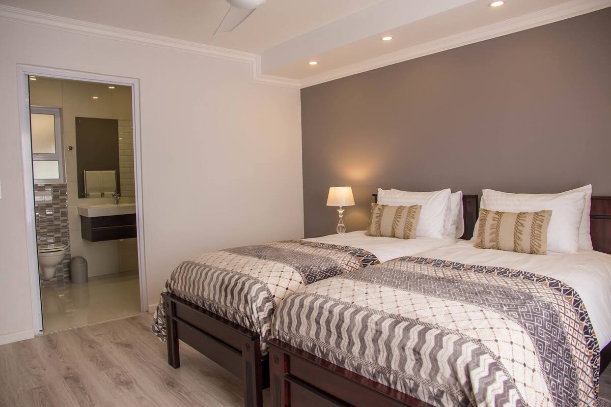 Un dormitorio con dos camas, un baño visible a través de una puerta abierta y una mesita de noche con lámpara.