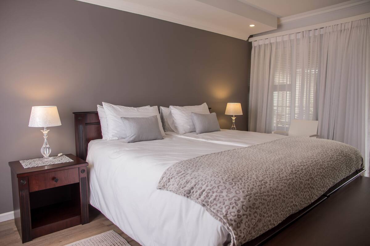 Un dormitorio con una cama grande, dos mesitas de noche con lámparas, un mueble auxiliar y una ventana con cortinas.