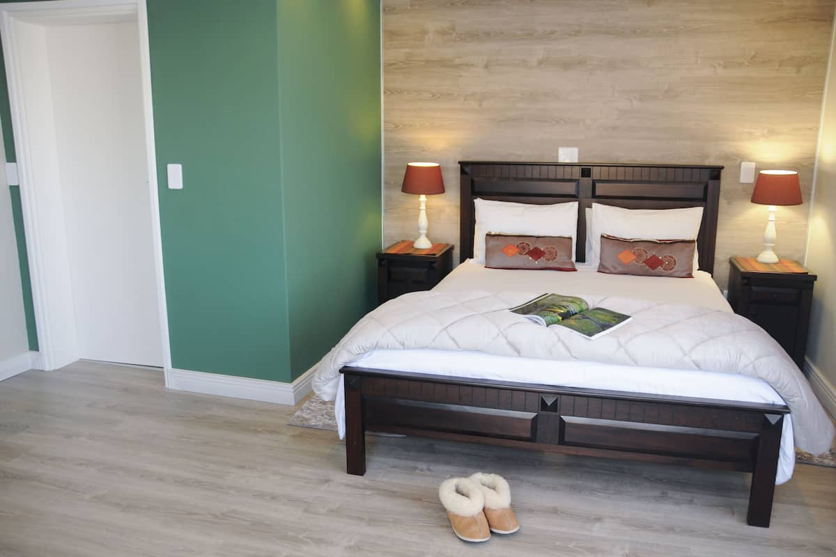Una cama bien hecha con un edredón acolchado y almohadas, dos lámparas de noche, un cabecero de madera y un par de pantuflas en el suelo.
