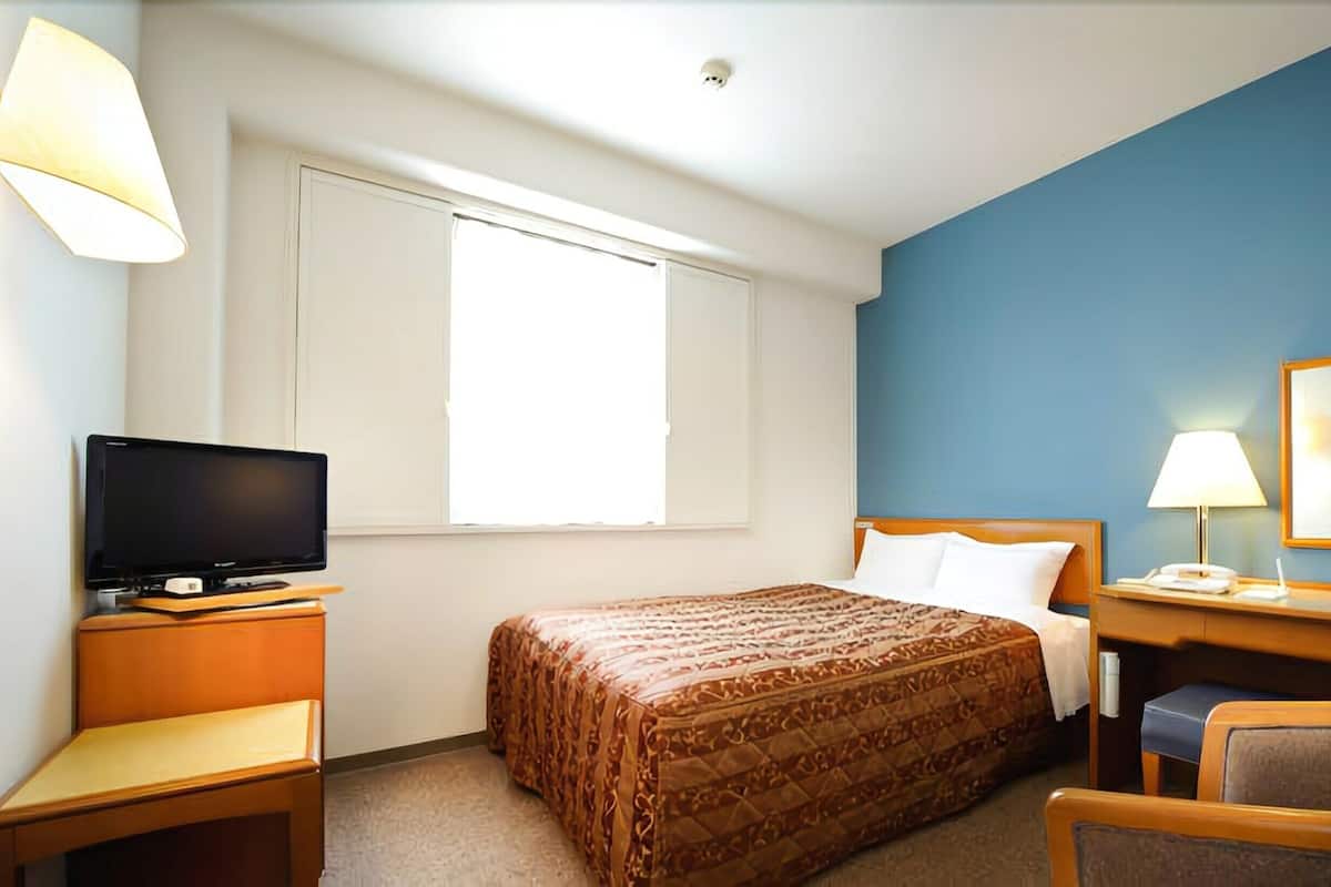 A hotel room with a bed, a desk with a lamp, a chair, a television, and a window.