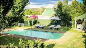 Ein Pool mit Liegestühlen und einer überdachten Sitzgelegenheit, umgeben von viel Grün und einem Haus mit Gründach.