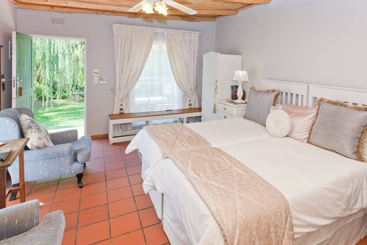 Ein Schlafzimmer mit Bett, Sessel, Schreibtisch und einem Fenster mit Vorhängen sowie Blick auf einen Garten.