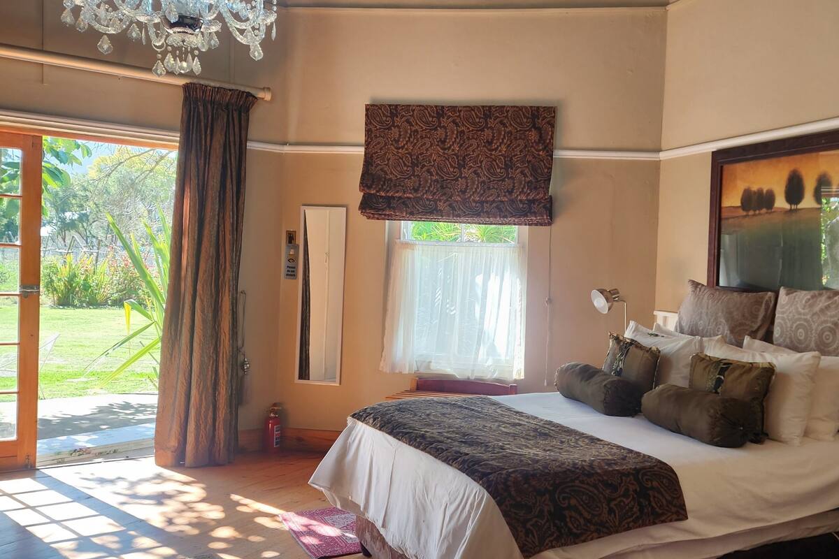 Ein Schlafzimmer mit einem großen Bett, einer Kristallleuchtre, einem Fenster mit Vorhängen und Blick ins Grüne.