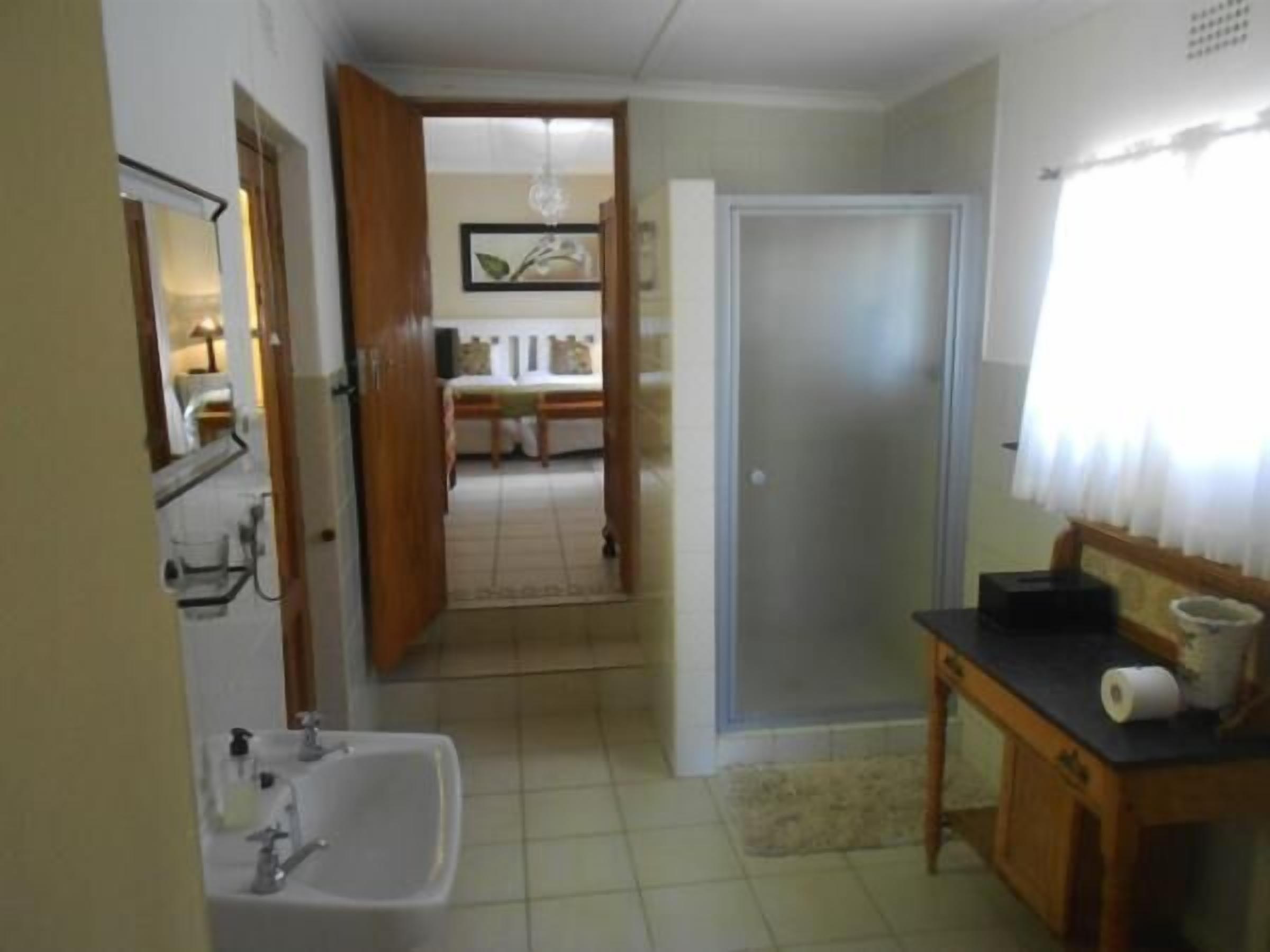 A bathroom with a sink, mirror, and toilet. There is a wooden cabinet with a black countertop and a roll of toilet paper. A door leads to a bedroom with a bed and a nightstand.