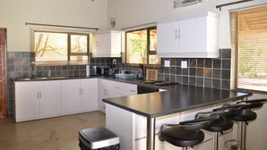 A kitchen with white cabinets, a central island with bar stools, a built-in stove, and a window with blinds. - Raptors Wildlife Escapes (Hoedspruit)