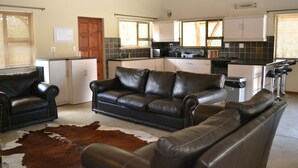 A modern kitchen with white cabinets, a black countertop, and a built-in stove. There are two black leather sofas and a cowhide rug in the living area. - Raptors Wildlife Escapes (Hoedspruit)