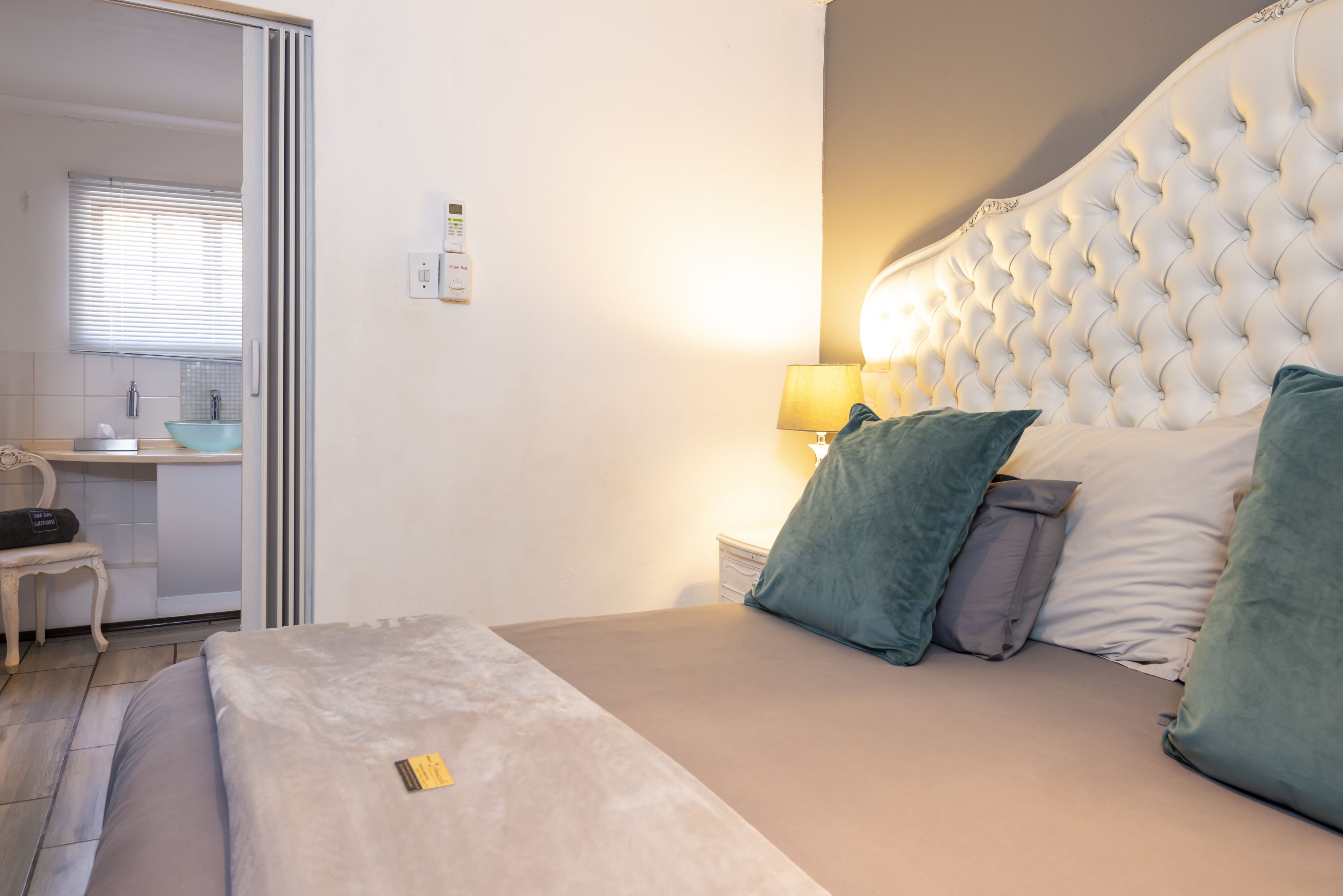 A bedroom with a large bed, a bedside table with a lamp, and a desk with a chair in the background.