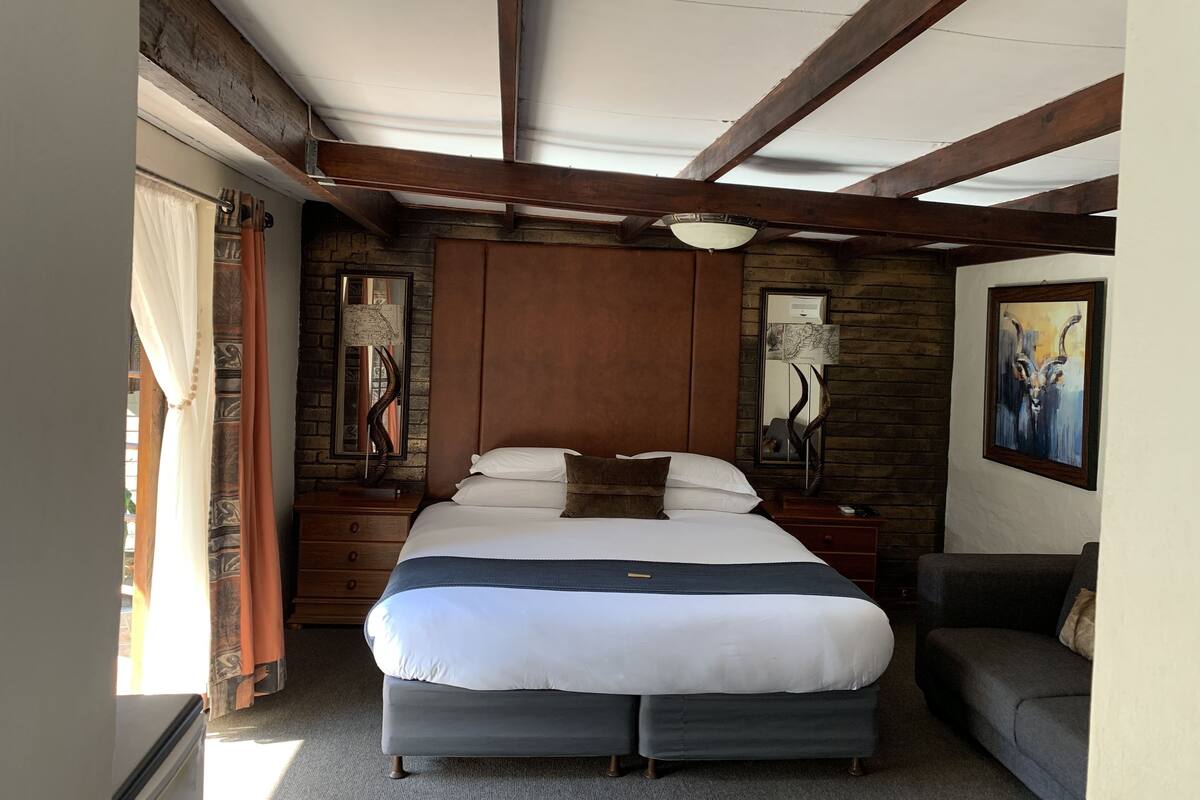 A bedroom with a large bed, wooden beams, a dresser, a nightstand, a lamp, and a painting on the wall.