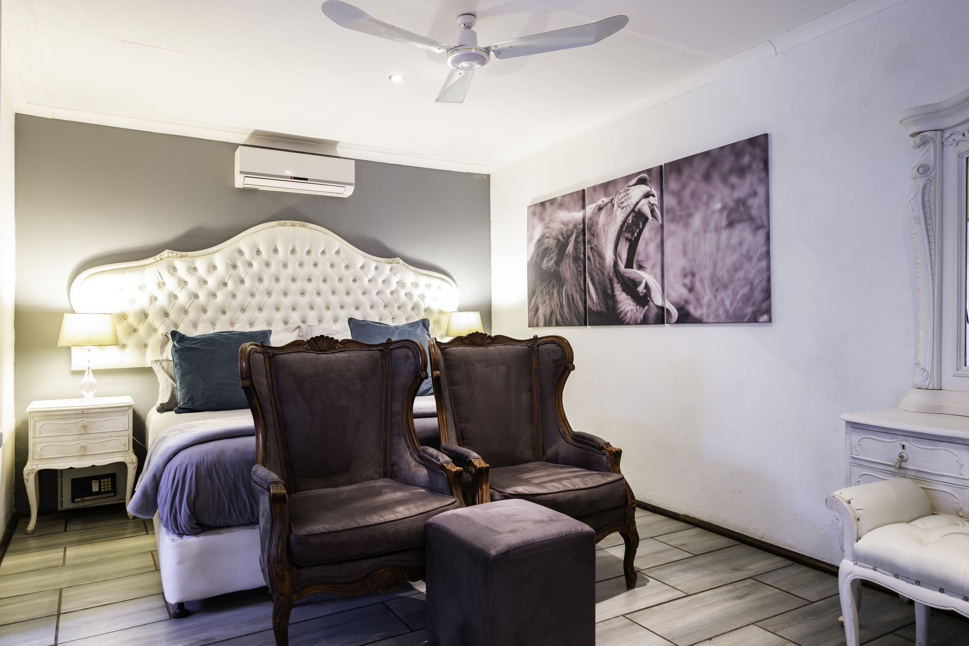 A bedroom with a tufted headboard, two armchairs, a bedside table with a lamp, and a large abstract painting on the wall.