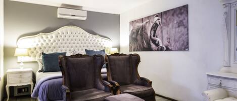 A bedroom with a tufted headboard, two armchairs, a bedside table with a lamp, and a large abstract painting on the wall.