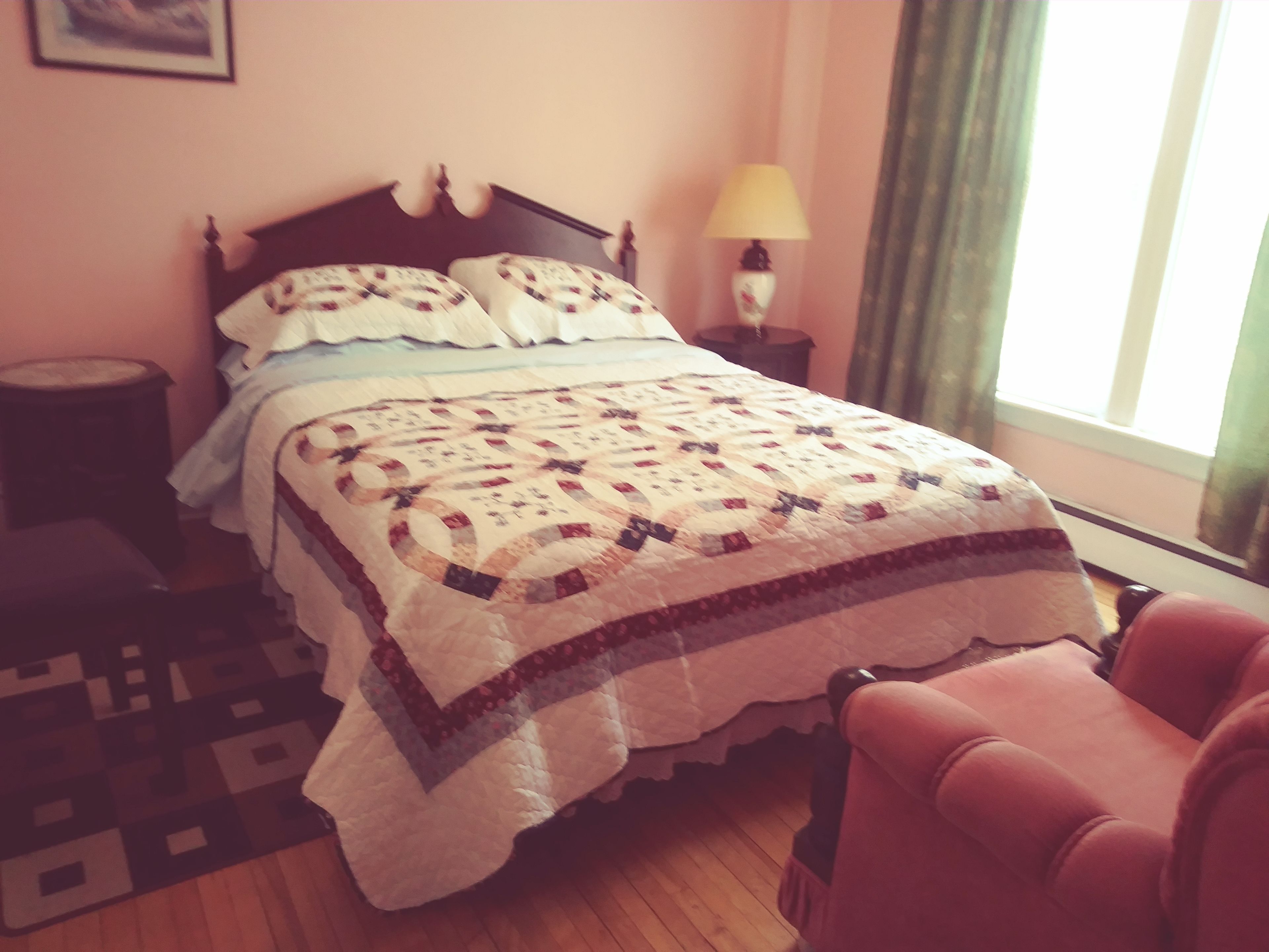 Una cama bien tendida con un edredón estampado, una lámpara de noche y una silla en una habitación con piso de madera.