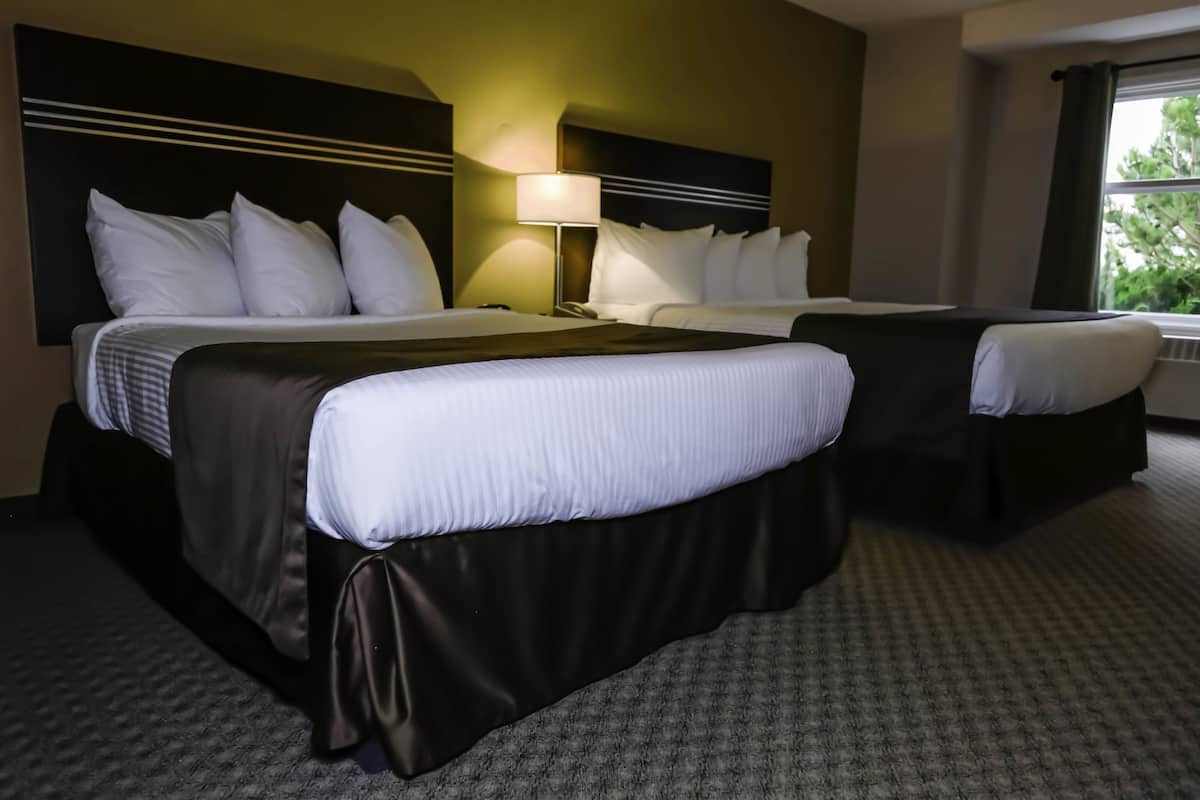 A hotel room with two beds, a nightstand, a lamp, and a window with a view of greenery.