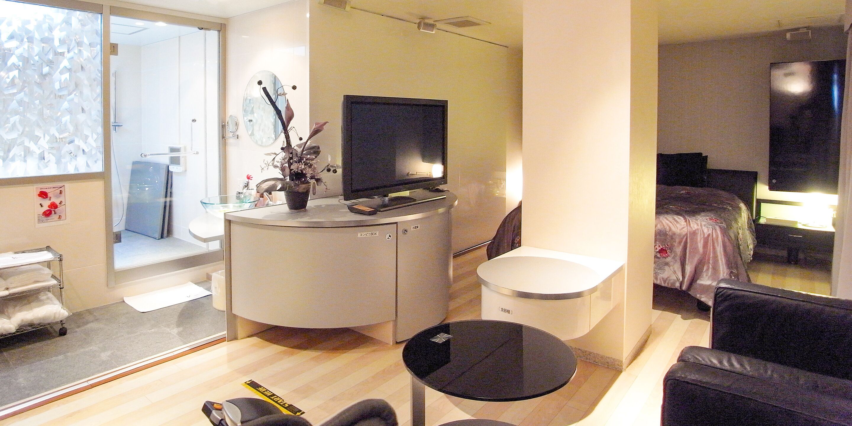 A modern hotel room with a flat-screen TV, a small round table, a sofa, a bed, and a bathroom with a glass shower enclosure.