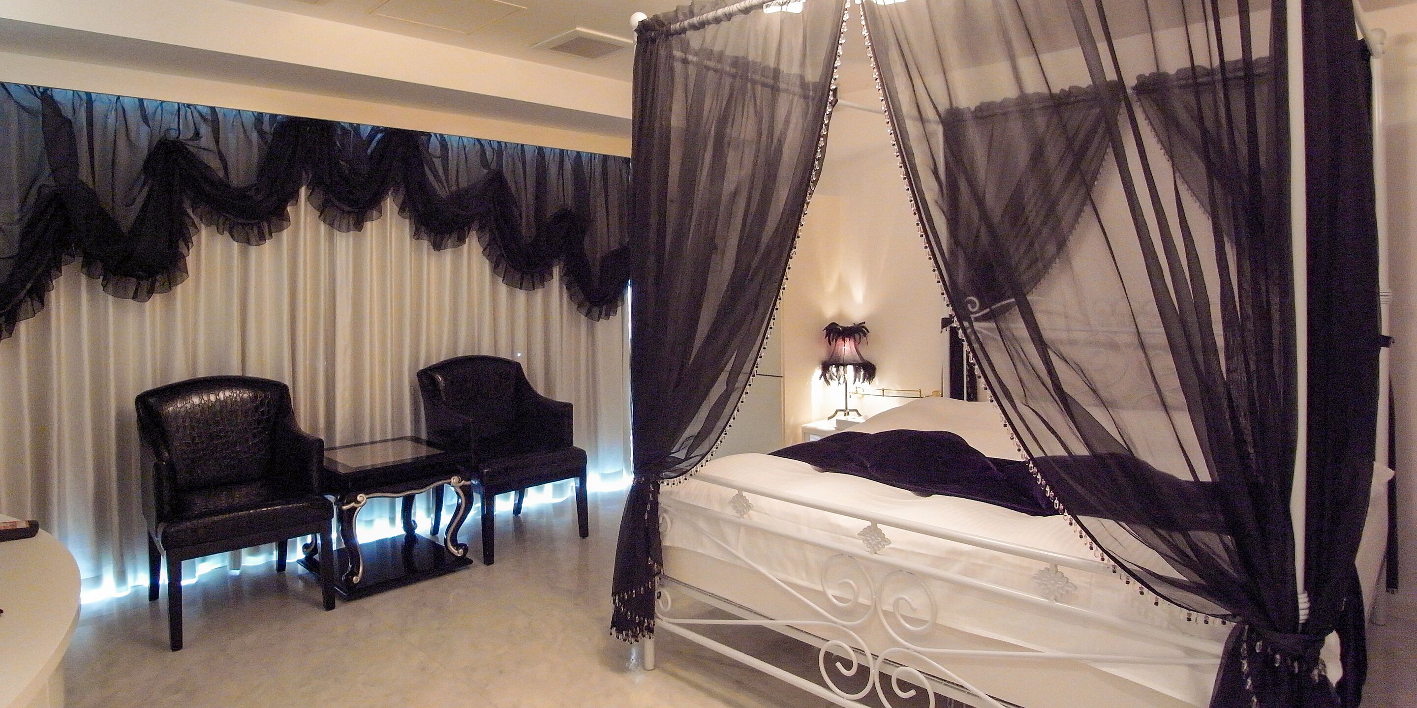 A bedroom with a four-poster bed, a nightstand, two chairs, and a large window with sheer curtains.