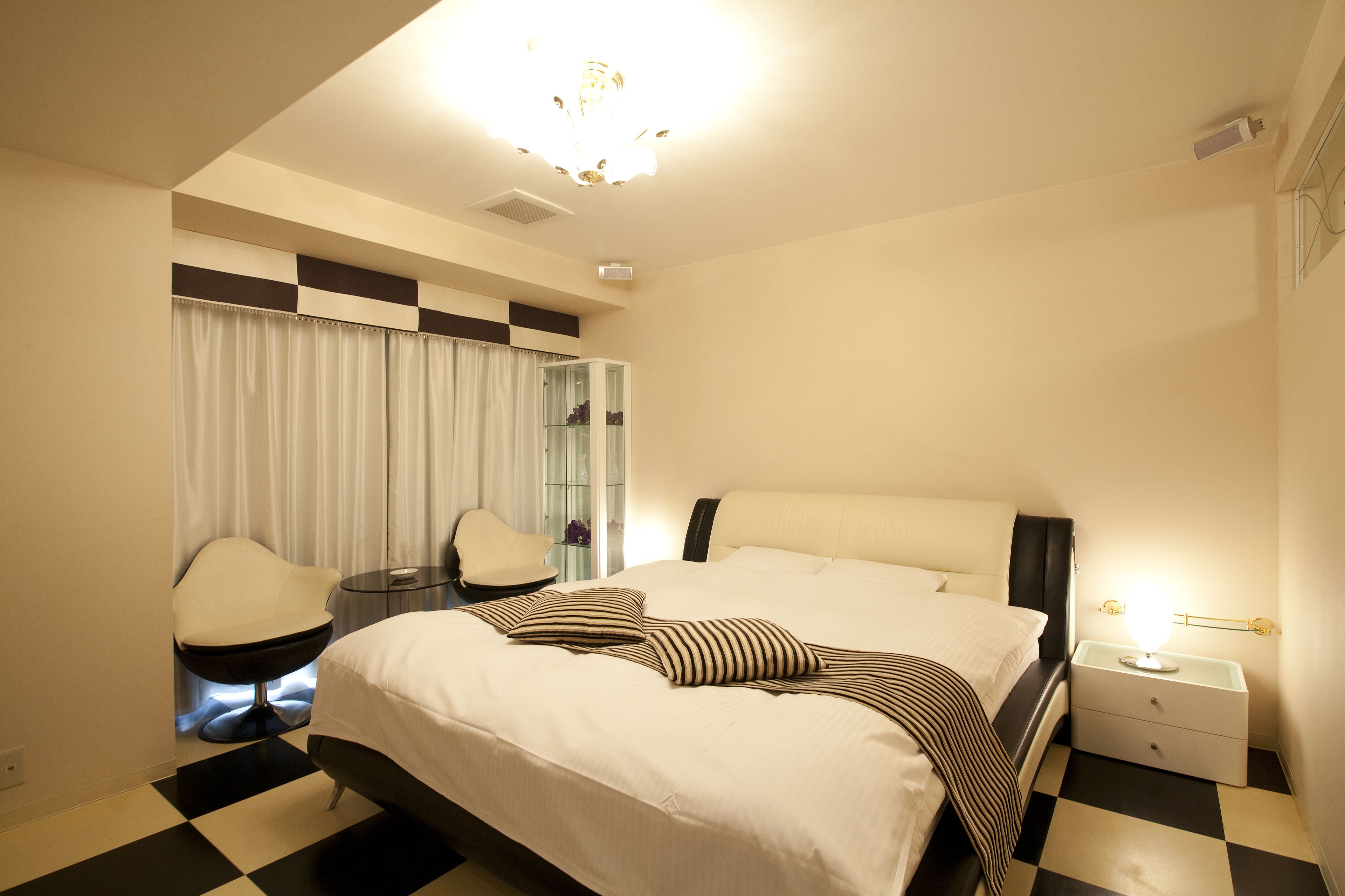 A modern hotel room with a large bed, a sitting area with a chair, a small table, and a nightstand with a lamp.