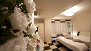 Una camera d'albergo con pavimento a scacchi, un letto, una sedia e un vaso con fiori bianchi.