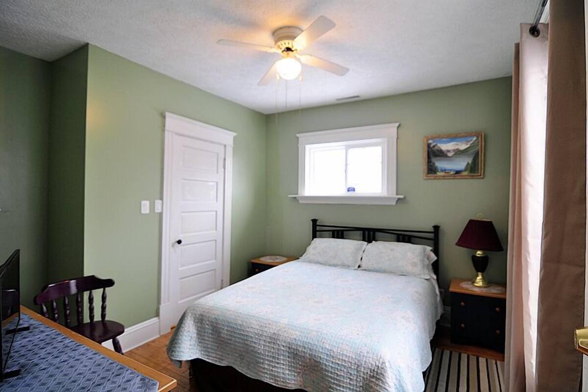 A bedroom with a bed, a window, a ceiling fan, a nightstand, and a chair.