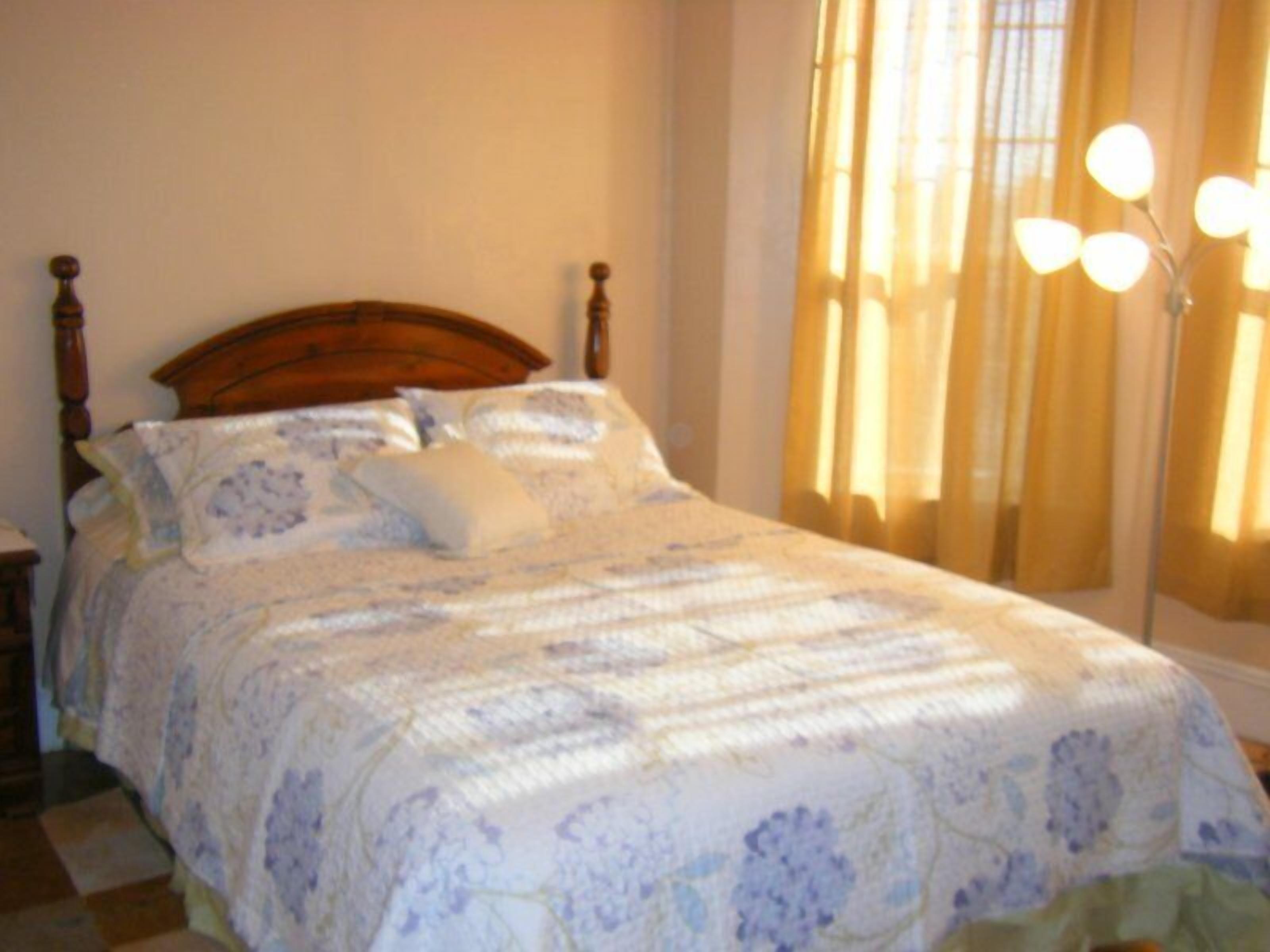 A neatly made bed with a floral-patterned bedspread and pillows, a wooden headboard, and a nightstand with a lamp.