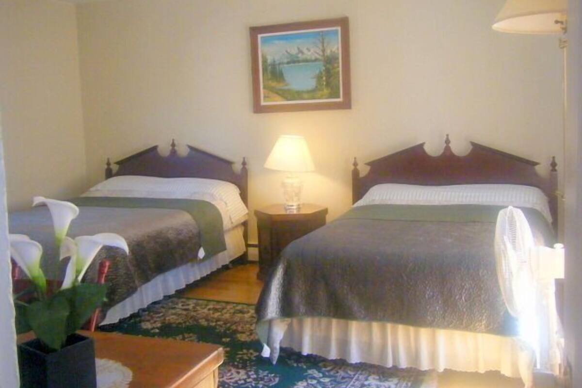 A hotel room with two single beds, a nightstand with a lamp, a framed picture on the wall, and a potted plant.