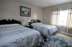 Un dormitorio con dos camas, una mesita de noche con lámpara, un cuadro enmarcado en la pared y una ventana con cortinas.