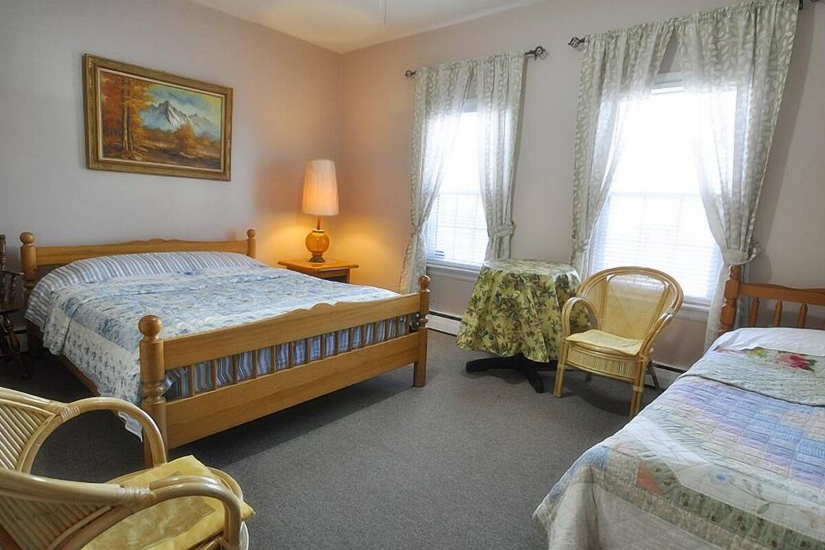 A bedroom with a wooden bed, a chair, a table with a lamp, and two windows with curtains.
