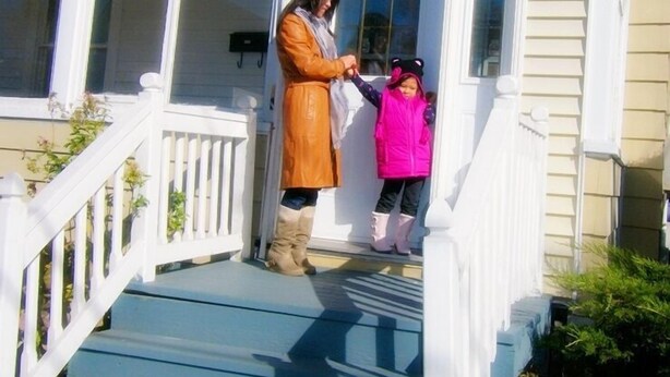 A woman in a brown coat and a child in a pink jacket standing on a porch with a white railing and a blue floor. - Glory Guest House (Moncton)