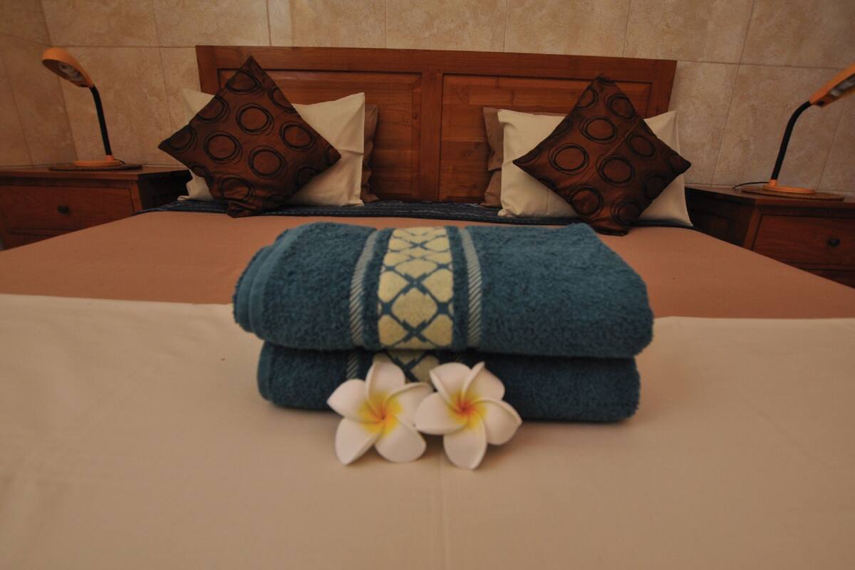 A neatly made bed with a blue and white patterned blanket, two white flowers, and two brown pillows with circular patterns.