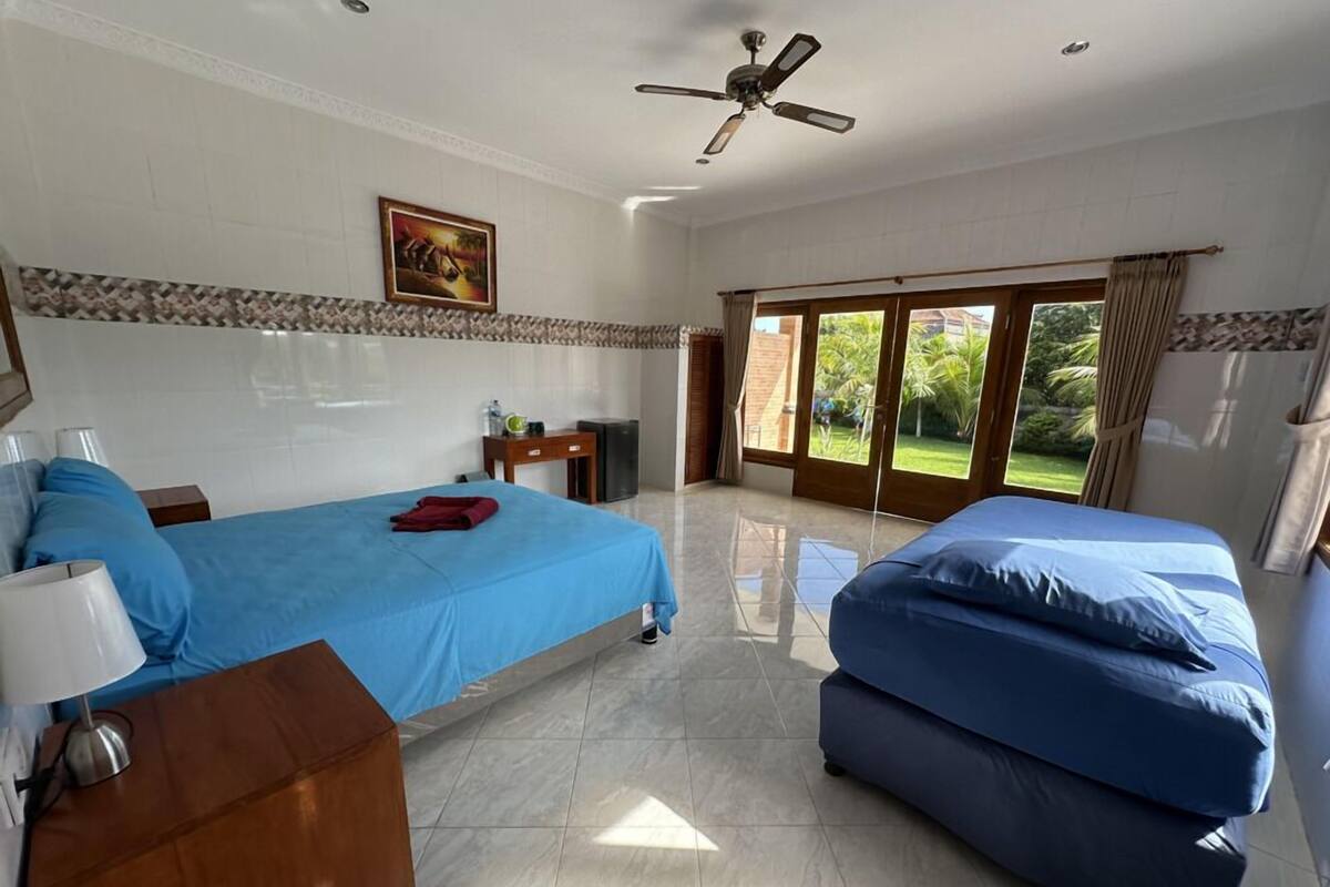 A hotel room with two beds, a ceiling fan, a painting on the wall, and a view of the outdoors through the door.