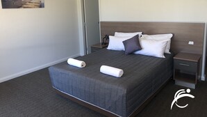 A neatly made bed with a gray quilt, two white pillows, and a rolled white towel. There is a small bedside table with a lamp, a wall-mounted air conditioner, and a door to the left. - Coro Club Motel (Griffith)